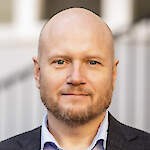 A calm, approachable man in a dark blazer and light blue shirt, photographed in soft outdoor light.