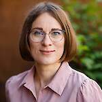 A woman in a pink shirt, with straight, brown, chin-length hair, wearing thin-framed glasses.