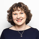 A woman with short, curly brown hair and blue eyes smiles warmly at the camera. She is wearing a dark blue top and a silver necklace with a flower-shaped pendant. The background is plain white.