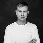 A black-and-white portrait of a clean-cut man with short hair, wearing a white T-shirt and a calm, slight smile with his arms crossed.